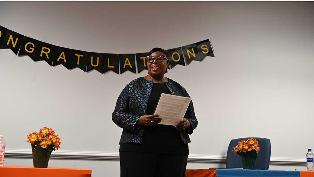 Deborah stands to speak holding her notes in a decorated conference room.