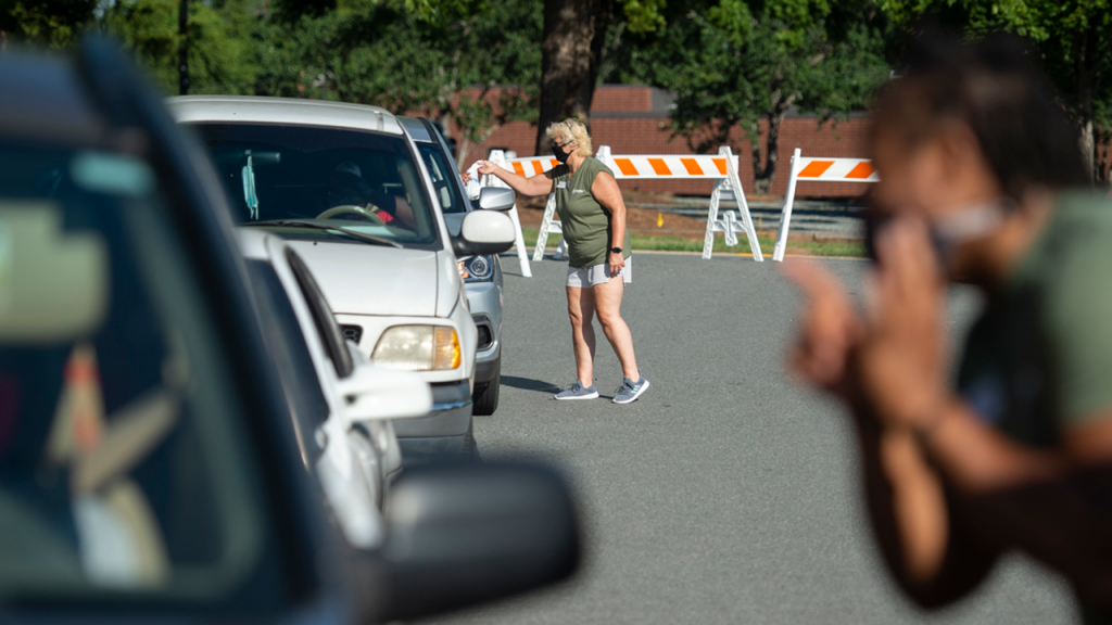 Volunteers wearing face coverings interact with individuals in cars lined up for services.
