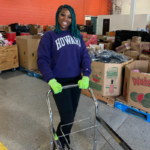 Howard University student in the warehouse, smiling, wearing green contruction gloves, pushing a cart.