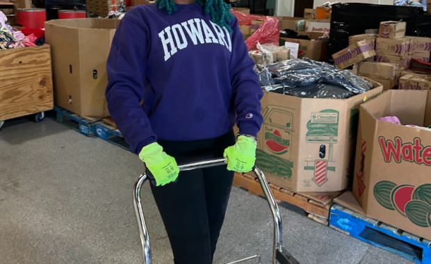 Howard University student in the warehouse, smiling, wearing green contruction gloves, pushing a cart.