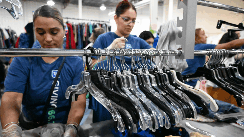 Two volunteer women wearing blue company shirts sorting clothes in the production warehouse behing racks with hangers on them.