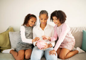 Black Mother And Daughters Holding Piggybank Putting Coins Sitting Indoors